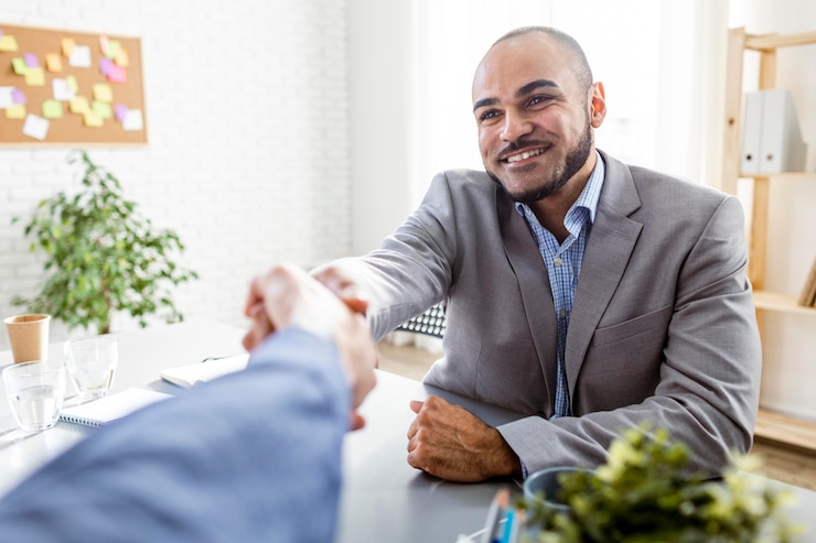 A smiling man in a grey suit shaking hands across a desk