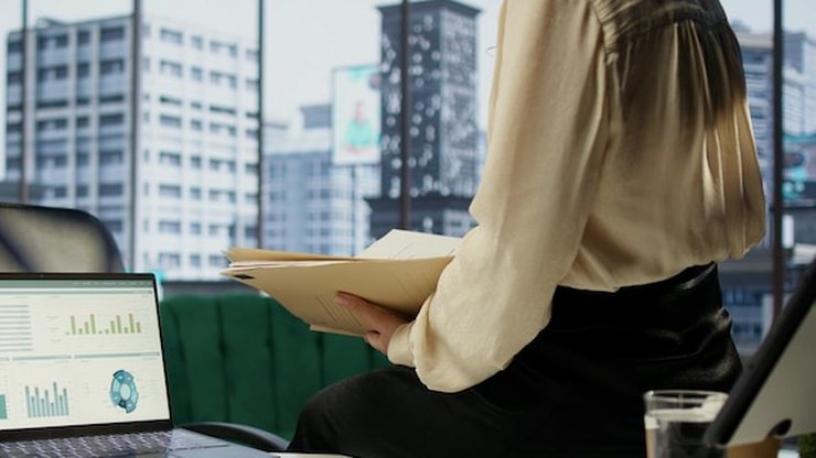 A woman holding a tablet beside a laptop showing charts by a city window