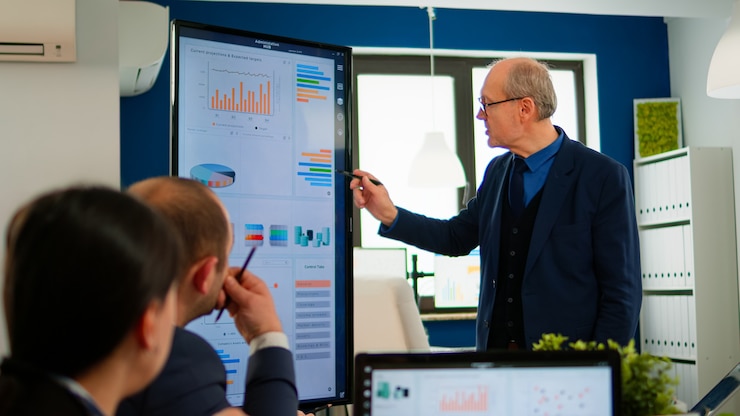 A man presenting a chart on a large wall screen to a small group